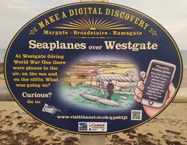 Blue oval metal sign on seafront railings with yellow and cream writing. Cartoon image of hand holding phone and man stood by vintage plane