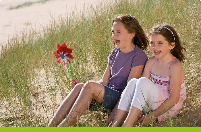 Two children sat in sand dunes, one holding a windmill with green stripe at bottom of image