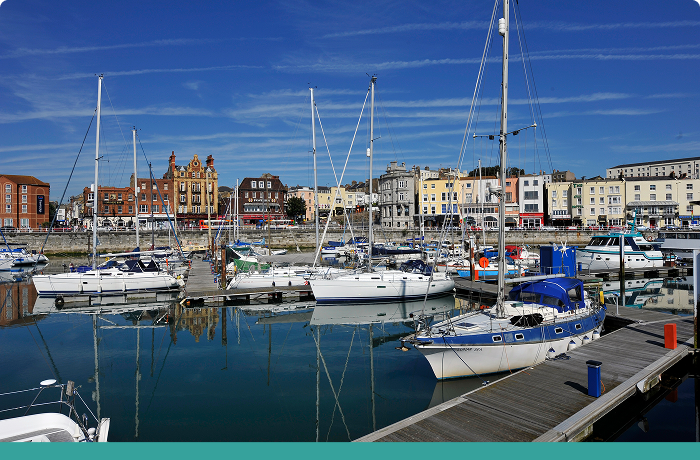 Yachts with sails down moored in harbour with shops/restaurants in background. Image with teal stripe at bottom