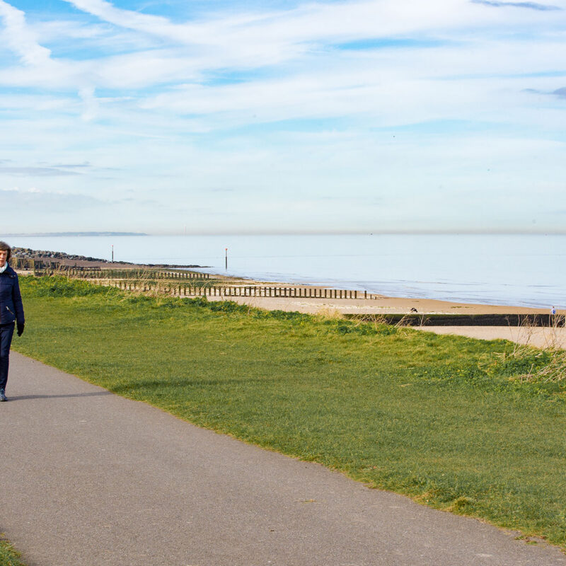 Couple walking along footpath by grass above sandy beach. Fort towers in background