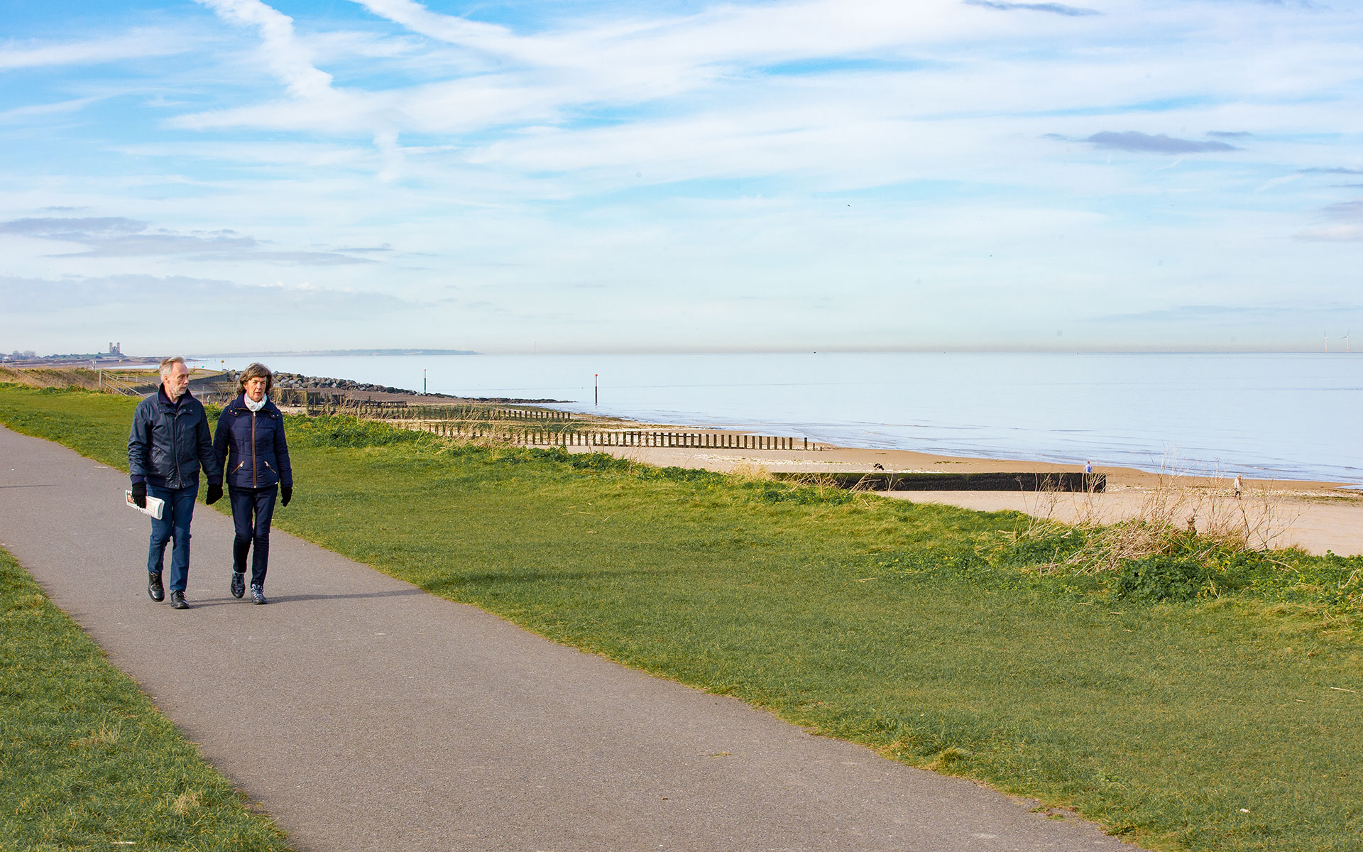 Couple walking along footpath by grass above sandy beach. Fort towers in background