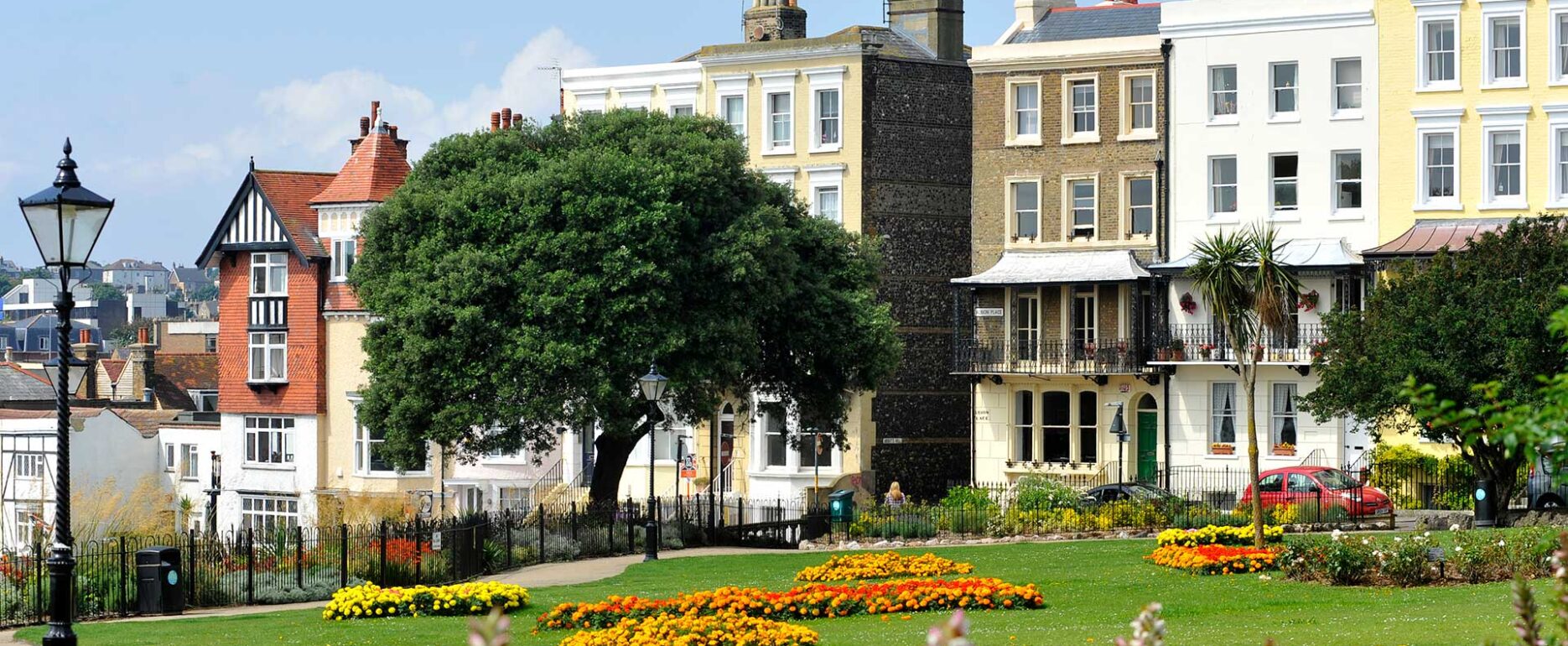 Park, garden area with buildings in background. Trees, lamp post and orange/yellow flowerbeds