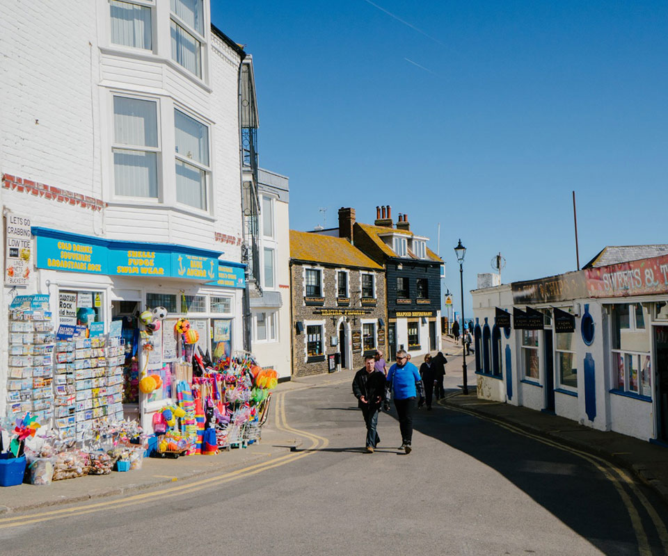 People walking on road between shops one with bucket, spades and postcards outside.