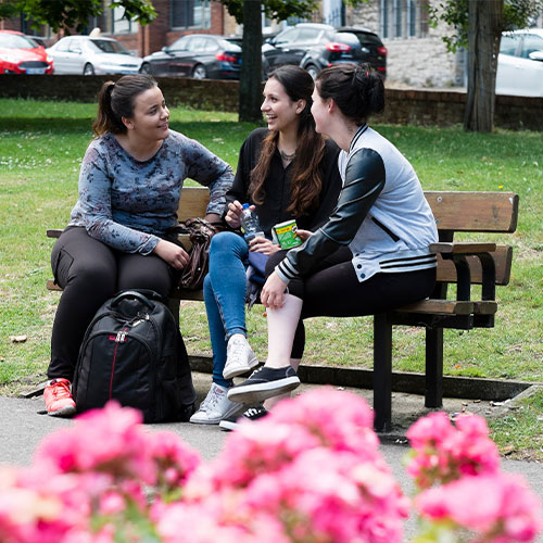 Three females sat on bench in park talking and eating