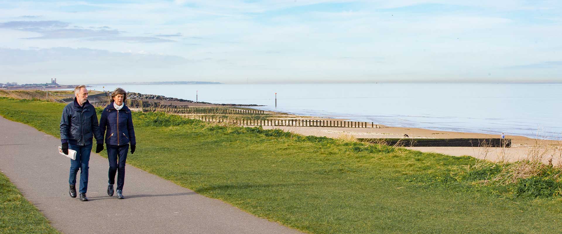 Couple walking along footpath by grass above sandy beach. Fort towers in background