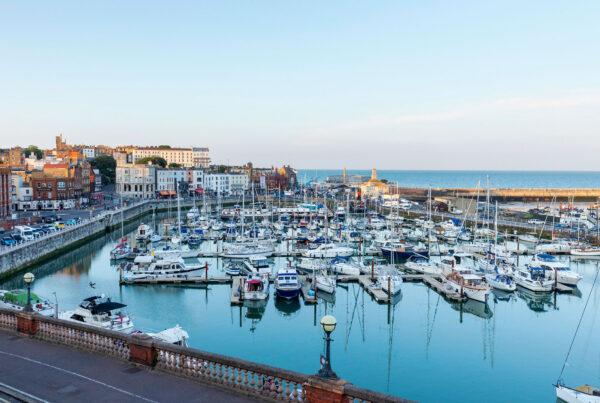 Ramsgate Royal Harbour during the day, featuring rows of white yachts and sailboats moored in the blue water, bordered by a stone promenade and historic buildings under a clear sky