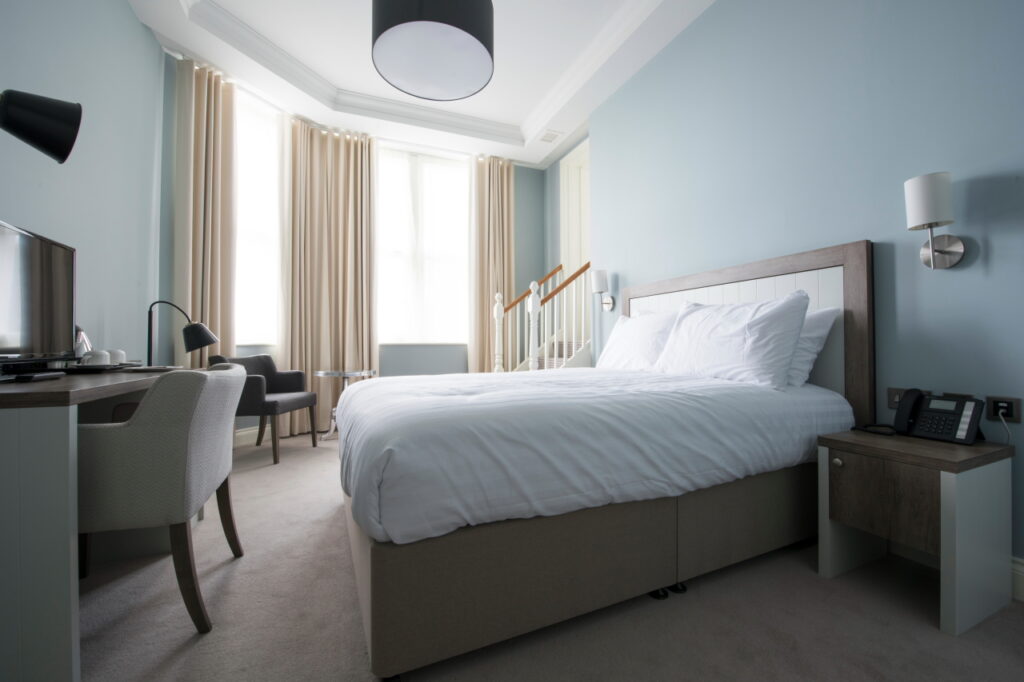 Double bed in centre of room with desk and chair at end. Large windows in background with chair and table. Stairs leading to bathroom