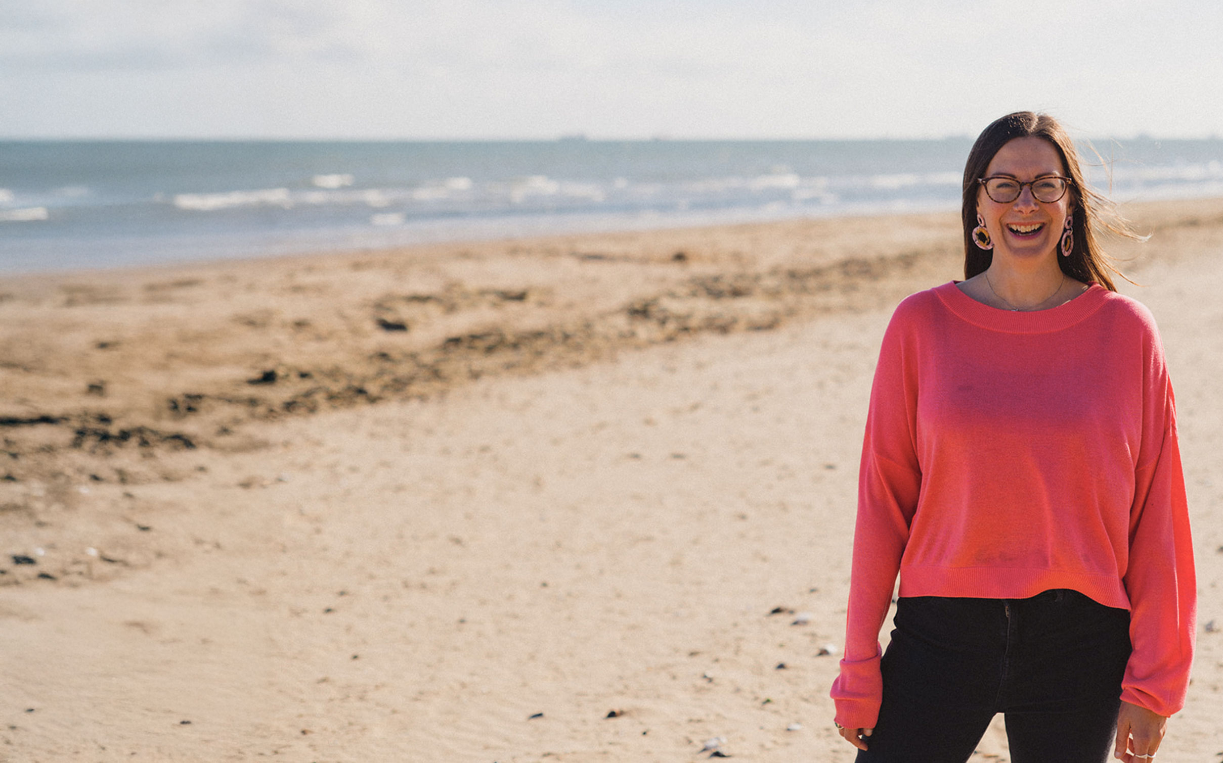 Lady in pink jumper stood smiling on sandy beach