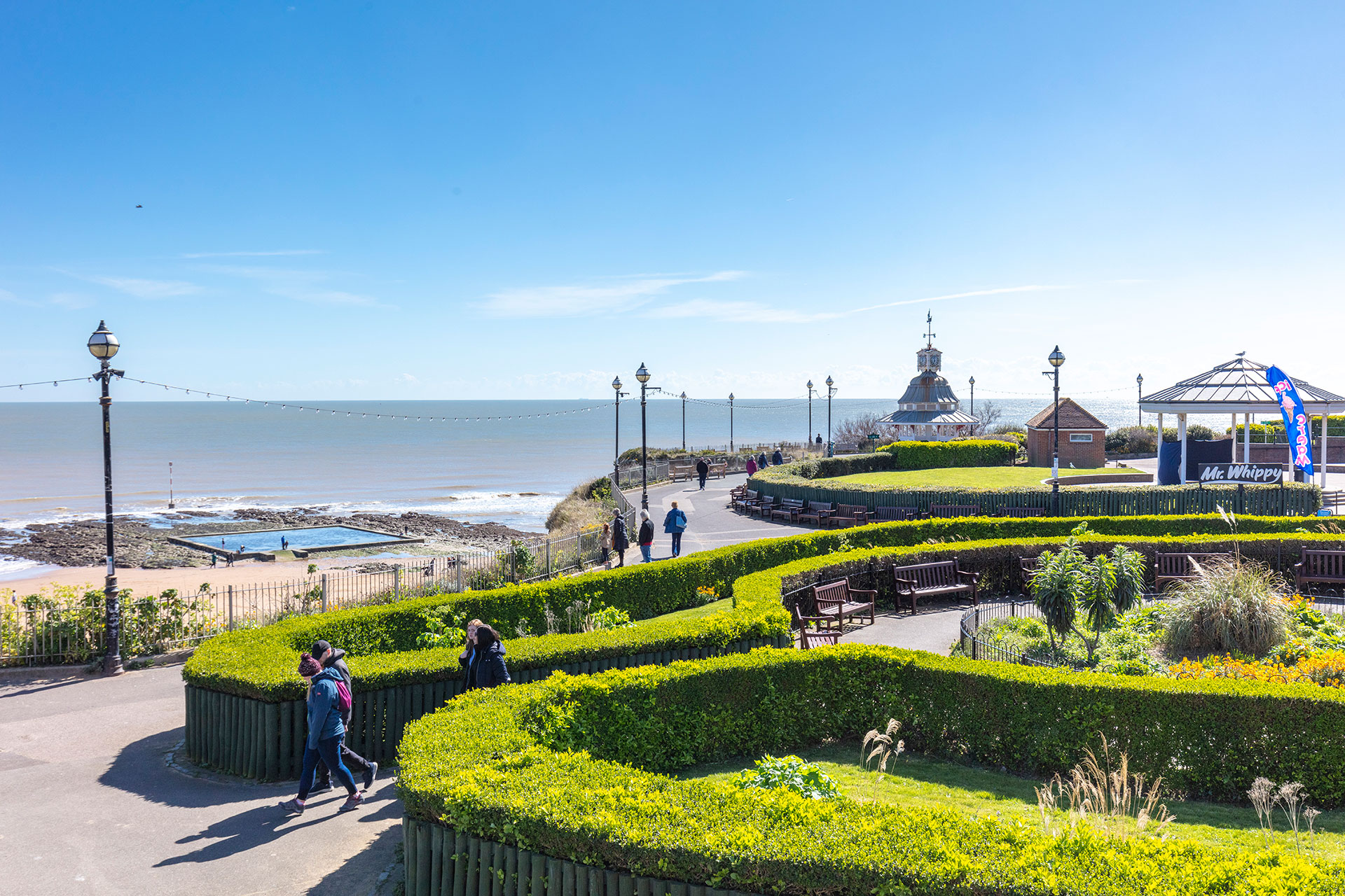 A sunny, high-angle view of Victoria Gardens in Broadstairs. Pedestrians walk along winding paths lined with manicured hedges and wooden benches, overlooking the coast and a small tidal pool on the beach below.