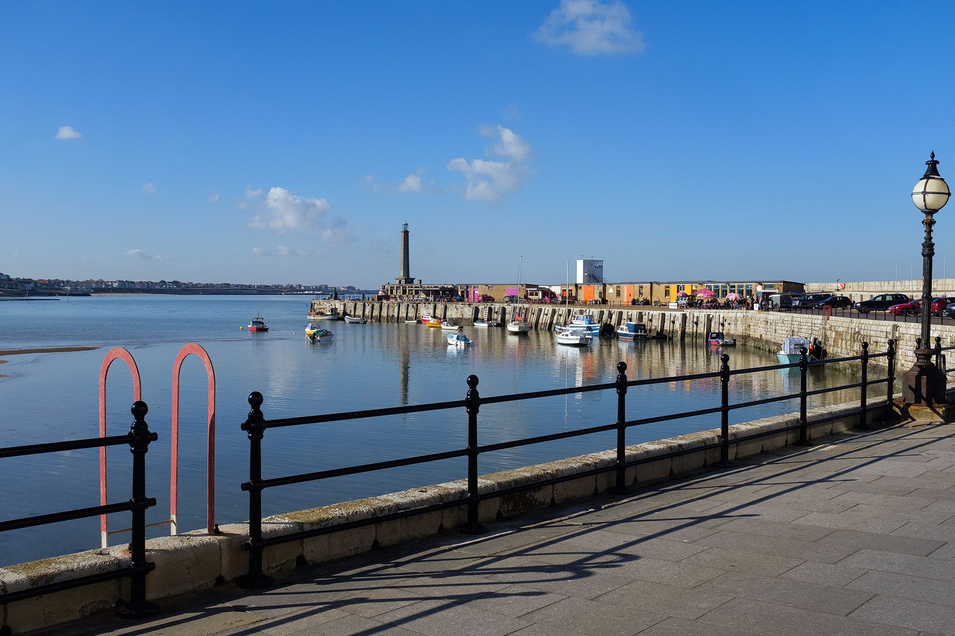 A view of the Margate Harbour Arm on a clear day, as seen from the stone pier. The calm blue water is dotted with small fishing boats, and colorful storage containers and stalls line the arm leading toward the lighthouse in the distance.