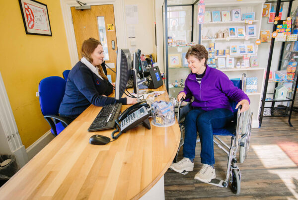 A staff member at a Visitor Information Centre sits behind a wooden desk, smiling while assisting a woman in a wheelchair. The office features yellow walls and shelves stocked with local cards and brochures.