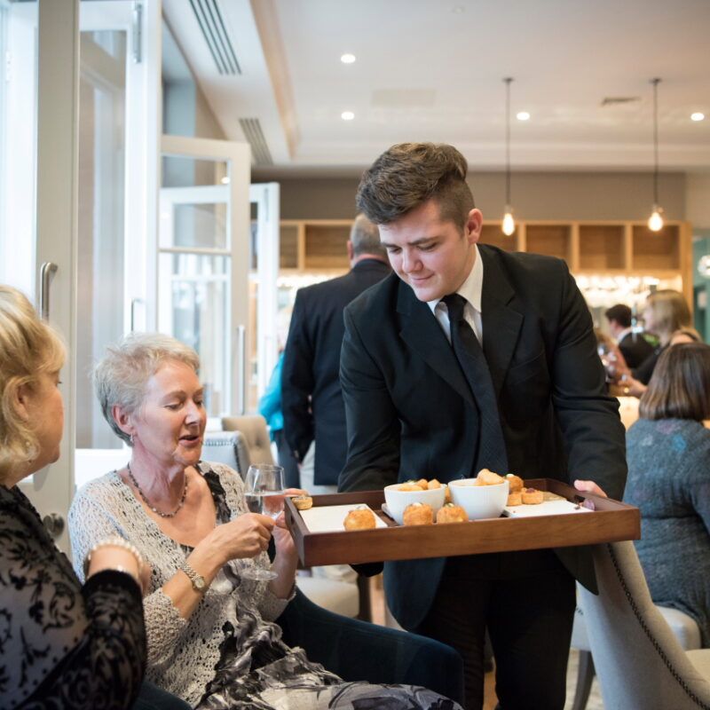 A young male server in a black suit and tie presents a wooden tray of canapés to two women seated in a bright, modern hotel bar area at The Yarrow Hotel in Broadstairs.