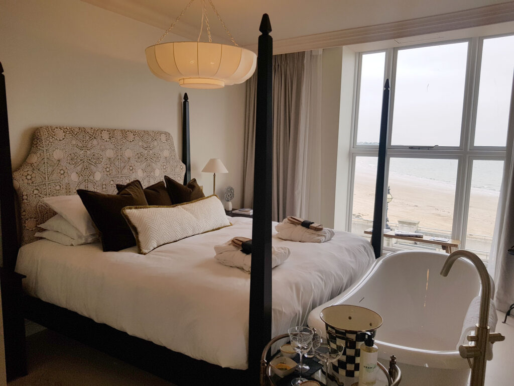 A luxury hotel suite at No.42 by GuestHouse Hotels in Margate, featuring a four-poster bed with a decorative headboard and a white freestanding bathtub positioned next to a large window overlooking the beach and sea.
