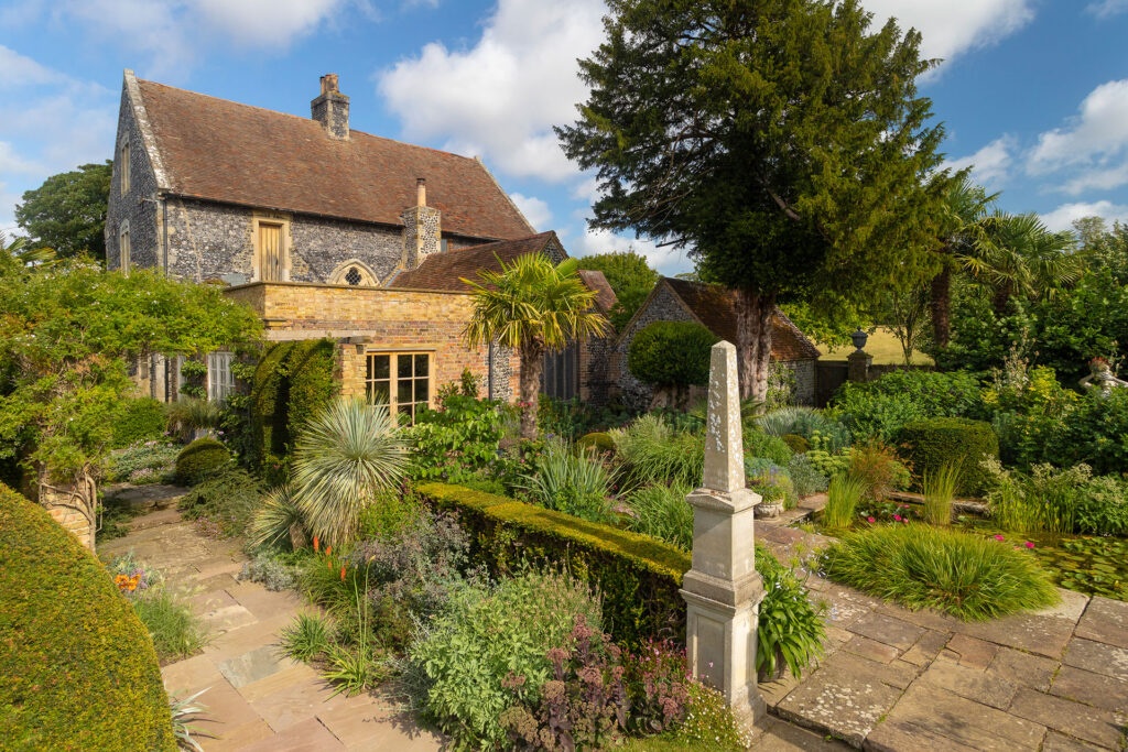 A beautiful stone and brick manor house at Chapel House Estate in Ramsgate, surrounded by a lush, manicured garden with a stone path, obelisk monument, and a small pond.