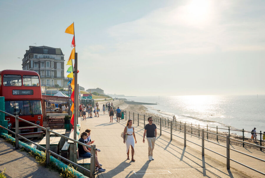 Pedestrians stroll along the sunny Margate promenade past The Sun Deck, which features a converted red double-decker bus, colorful flags, and outdoor seating overlooking the sandy beach and sea.