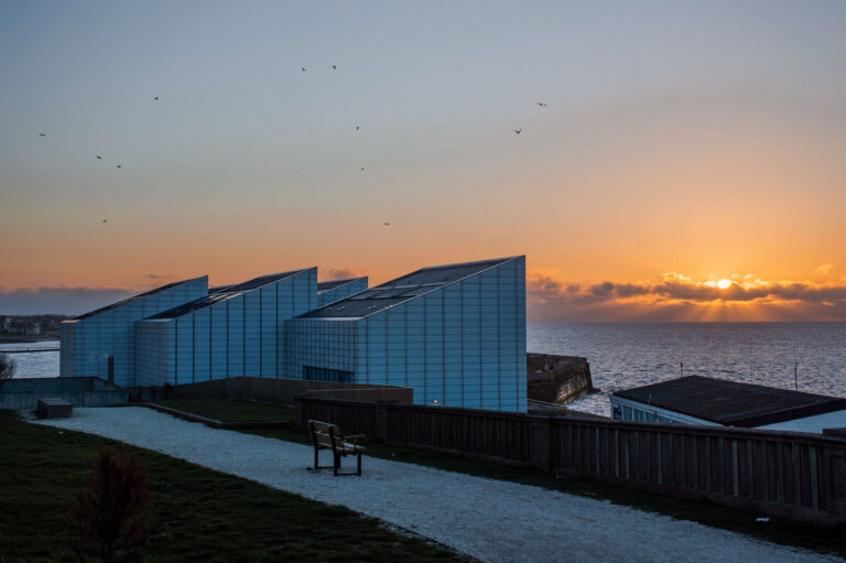 A dramatic sunset over the sea in Margate, featuring the modern, white angular architecture of the Turner Contemporary gallery. Birds fly across the orange and grey sky as the sun dips behind a line of low clouds.
