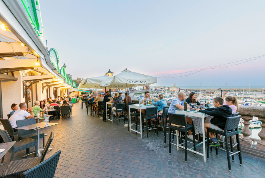 Patrons enjoy evening alfresco dining on the terrace of West Cliff Arcade in Ramsgate. The scene shows multiple groups of people at tables overlooking the Royal Harbour, which is filled with boats under a soft dusk sky.