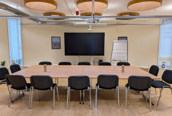 A boardroom-style meeting room featuring a large wooden table surrounded by black chairs. A large flat-screen monitor and a flipchart are positioned at the head of the table.