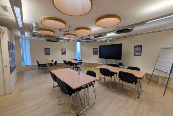 A classroom-style setup in a modern meeting room, featuring several light wooden tables with black chairs arranged for small group discussions. A large flat-screen monitor and a flipchart are positioned at the front of the room.