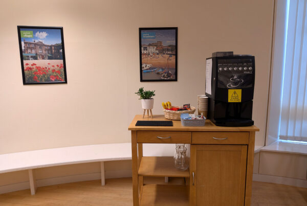 A self-service refreshment station in a modern meeting room, featuring a coffee machine, a basket of snacks, and disposable cups on a light wooden cabinet.