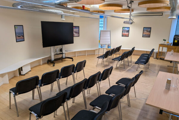 An empty theater-style training room with rows of black chairs facing a large mobile flat-screen monitor and a flipchart.