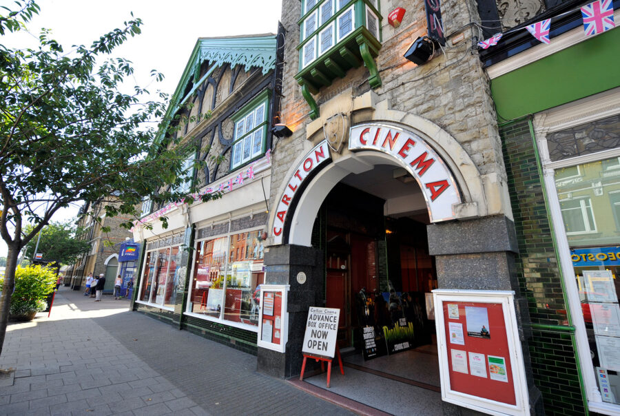 Arched entrance to cinema building, name above and sign welcoming. Shop windows to either side