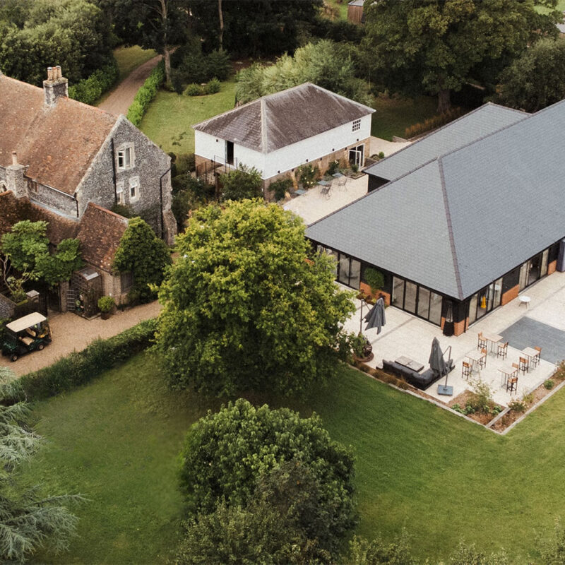 Aerial view of a historic stone building and a modern dark event space, connected by gardens and a paved terrace