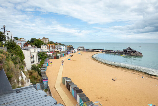 Aerial view of sandy beach looking towards pier arm with beach huts at back of beach, wooden boardwalk and tide half way in