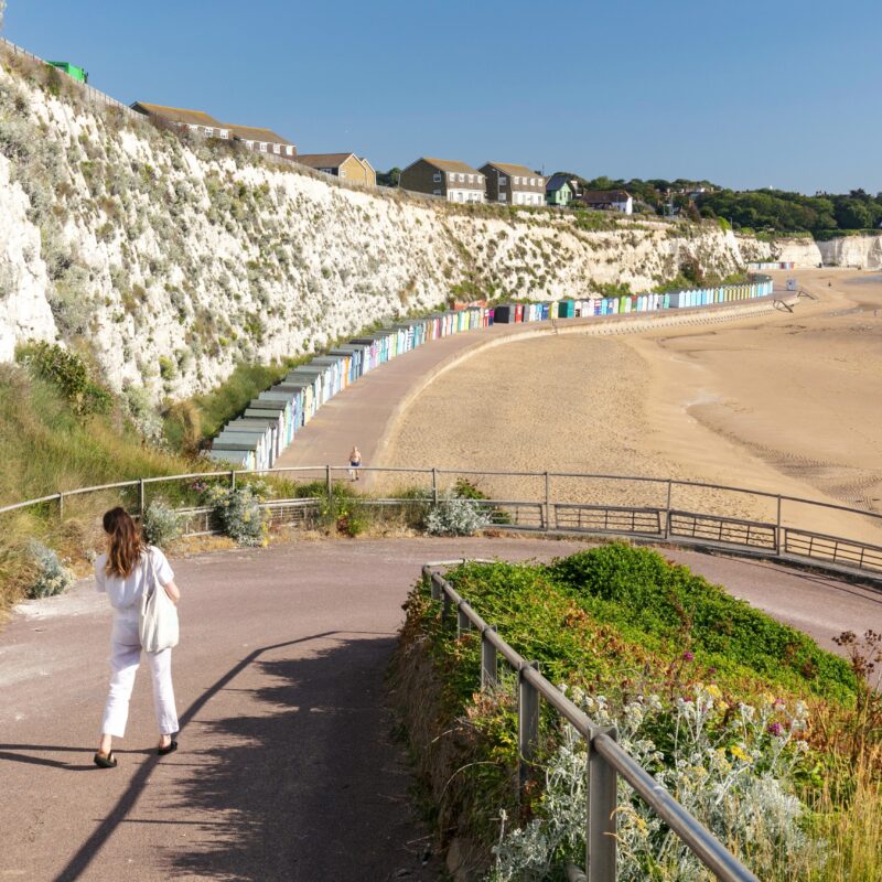 man and lady walking down ramp to sandy beach. Row of beach huts in distance
