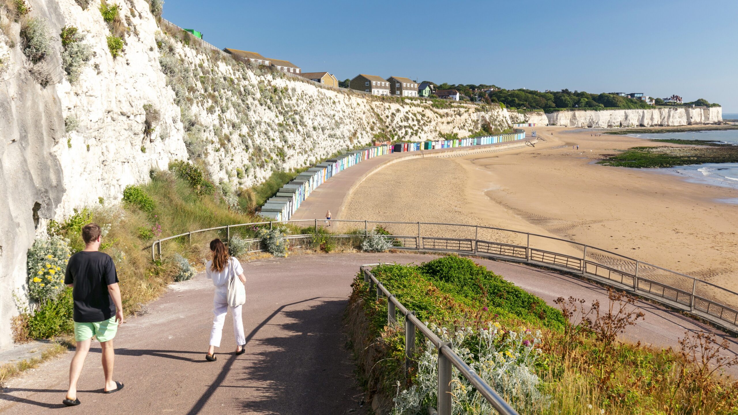 man and lady walking down ramp to sandy beach. Row of beach huts in distance